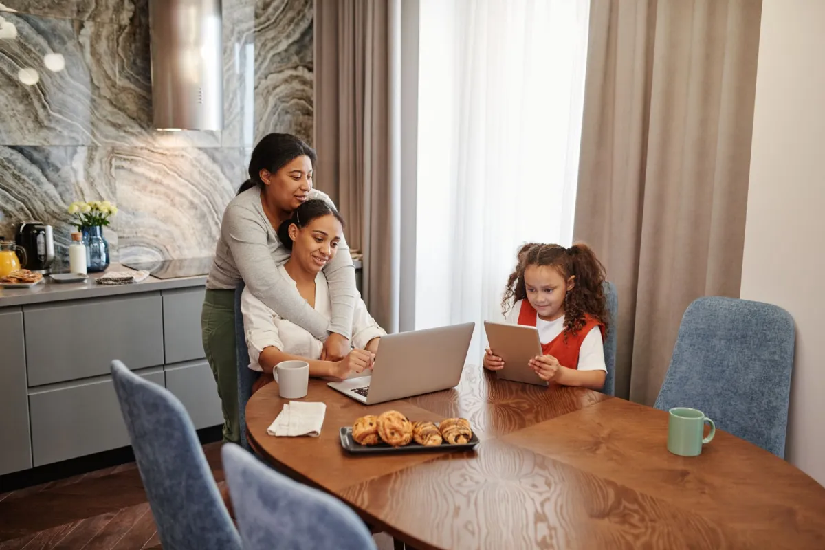 Two adults and a teenager sit at a kitchen table, each working on their own laptop.